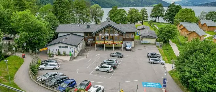 Drohnenaufnahme eines Parkplatzes der gut besucht ist, im Hintergrund sieht man Berge und einen See