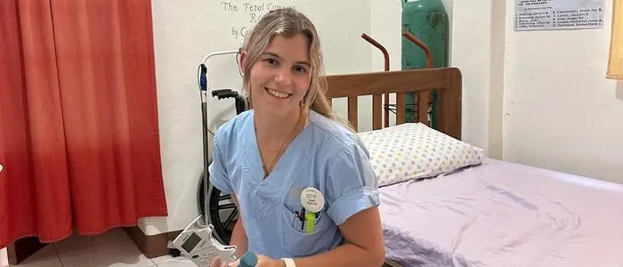 Eine junge Dame sitzt auf einem Medizinball. Sie hat einen blauen Krankenhaus-Kittel an und medizinische Geräte in den Händen