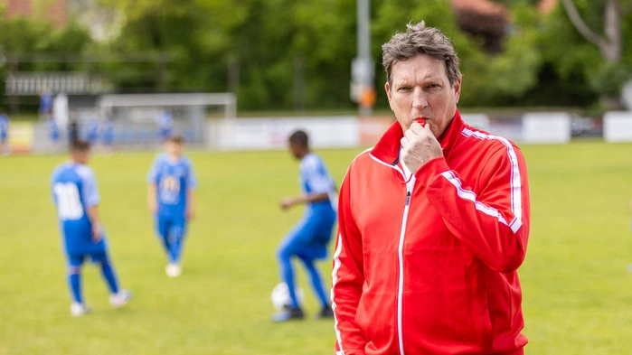 Fußballlegende Andi Herzog bläst in eine Trillerpfeife. Im Hintergrund trainieren drei Nachwuchsspieler