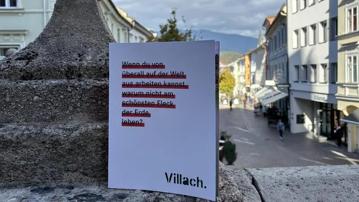 Eine Broschüre angelehnt an eine Mauer, im Hintergrund der Villacher Hauptplatz