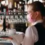 A female Waiter of European appearance in a medical mask serves Latte coffee