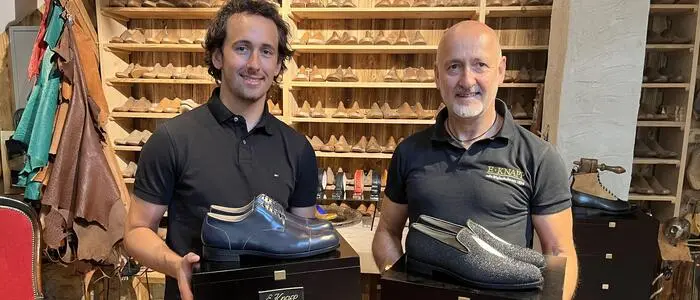 Zwei Männer, einer Mitte 50, einer Anfang 20, lächeln in die Kamera. Sie halten maßgefertigte Schuhe in den Händen.