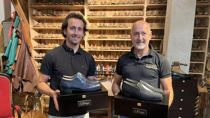 Zwei Männer, einer Mitte 50, einer Anfang 20, lächeln in die Kamera. Sie halten maßgefertigte Schuhe in den Händen.
