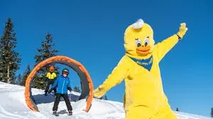 Bei strahlendem, blauem Himmel fahren zwei Kinder auf Skiern durch einen kleinen Tunnel, vor ihnen fährt ein Maskottchen als Ente | Vorfreude auch am Klippitztörl mit „Klippi Pippi“
