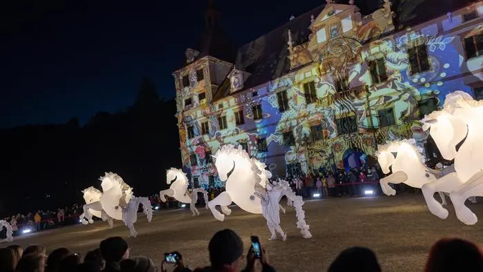 Lichter werden auf eine Schlossfassade projiziert. Im Vordergrund sind leuchtende Pferde aus Kunststoff zu sehen.