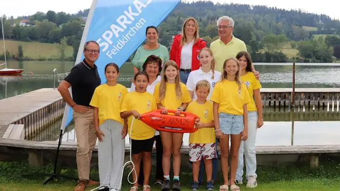 Mehrere Kinder in gelben Shirts sowie Erwachsene stehen vor einem See