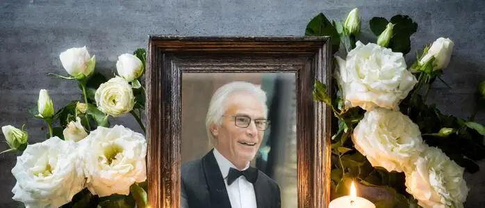 Gerahmtes Porträt eines grauhaarigen Mannes im Anzug mit Fliege, davor Kerzen und Blumen