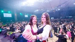 Zwei junge Frauen im Dirndl stehen auf einer Bühne, hinter ihnen tanzen Paare auf einer Tanzfläche