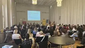 Blick von hinten auf einen vollen Saal mit Menschen, die sitzen und einem Vortrag lauschen