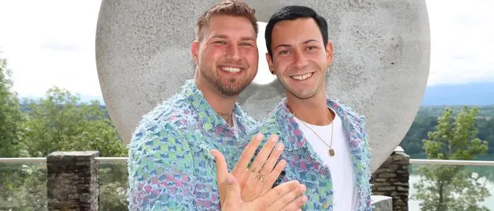 Zwei Männer im selben Outfit halten ihre Hände mit Ehering in die Kamera und lachen