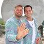Zwei Männer im selben Outfit halten ihre Hände mit Ehering in die Kamera und lachen