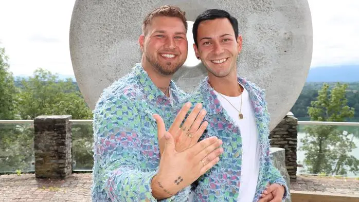 Zwei Männer im selben Outfit halten ihre Hände mit Ehering in die Kamera und lachen