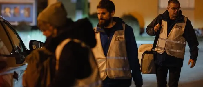 Drei Personen mit Warnwesten und gepackten Taschen gehen am Abend zu einem Auto.
