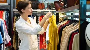 Young woman is choosing raincoat in clothing shop and holding one raincoat of mustard color. Photo with depth of field