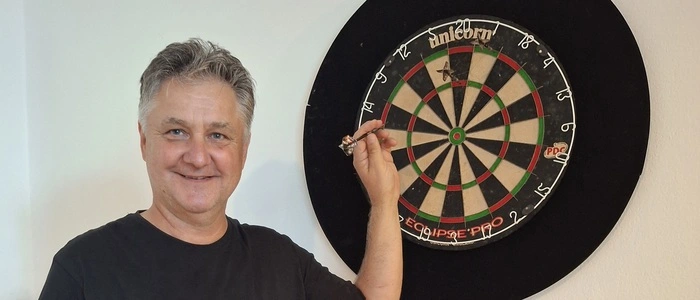 Ein Mann mit schwarzem T-Shirt steht vor einer Dartscheibe