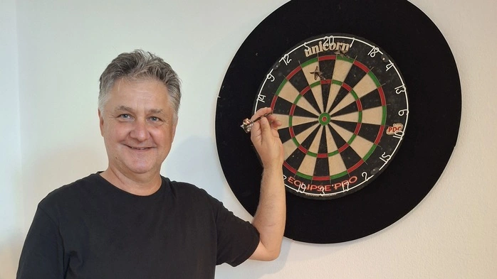 Ein Mann mit schwarzem T-Shirt steht vor einer Dartscheibe