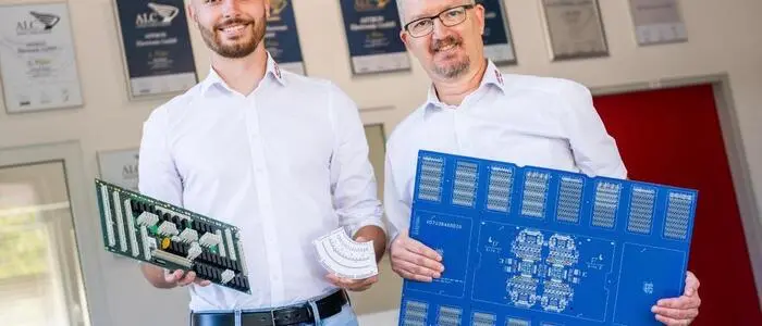 Zwei Männer stehen in einem Büro und halten elektronische Bauteile in den Händen und lächeln in die Kamera