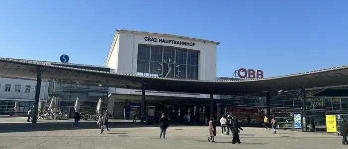 Das Bild zeigt den Grazer Hauptbahnhof von vorne. Einige Passanten halten sich im Vordergrund am Europaplatz auf.
