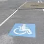 Ein asphaltierter Parkplatz vor einem Geschäft, am Boden ist eine rollstuhlfahrende Person als Kennzeichen für einen Behindertenparkplatz eingezeichnet