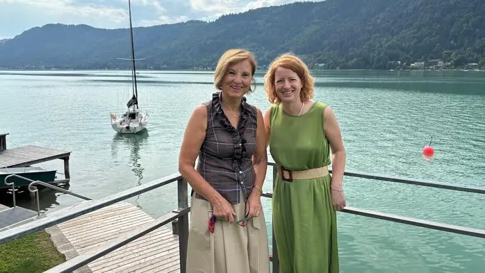 Zwei Damen stehen vor einem türkisblauen See, im Hintergrund ist ein Segelboot.