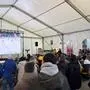 Blick in ein Zelt, darin stehen und sitzen zahlreiche Menschen und schauen auf die Leinwand