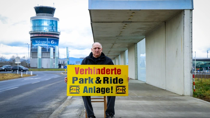 Adolf Egger sitzt mit einem großen gelben Schild mit der Aufschrift „Verhinderte Park & Ride Anlage!“ am Gelände vor dem Grazer Flughafen