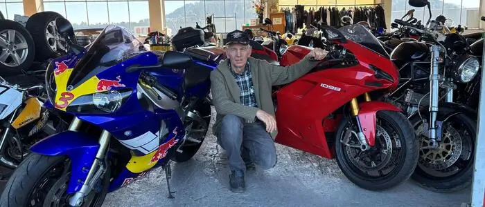 Ein älterer Herr hockt am Boden einer Werkstatt und ist von Motorrädern umgeben