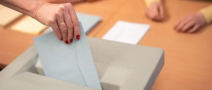 Eine Hand mit roten Fingernägeln wirft einen Brief in eine Wahlurne