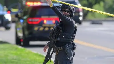 Schwer bewaffneter US-Polizist hebt ein Absperrband mit seinem linken Arm in die Höhe. Hinter ihm steht ein Einsatzwagen.
