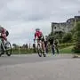 Mehrere Radfahrer im Dress und Radmontur radeln eine Straße entlang. Im Hintergrund sieht man eine Burg