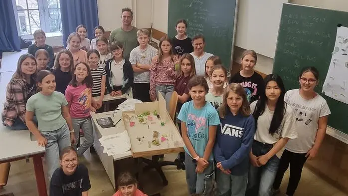 Eine Schulklasse hat sich im Klassenzimmer um ein Modell eines Gartens zusammengestellt und schaut in die Kamera