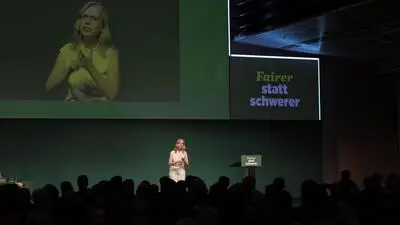 Leonore Gewessler auf der Bühne beim Grünen Bundeskongress; das Publikum als Silhouette im Vordergrund, Gewessler noch einmal groß auf der Leinwand im Hintergrund