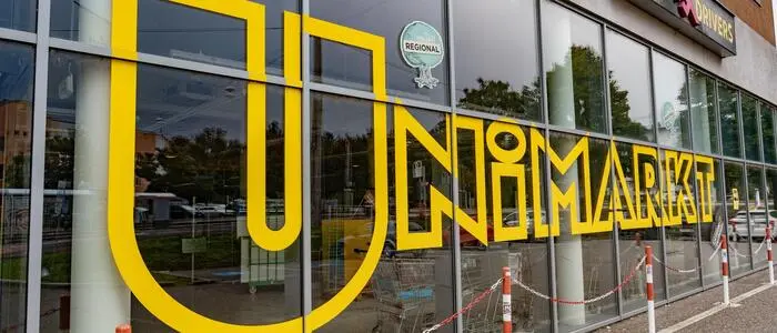 Auf dem Bild sieht man die Fassade eines Geschäfts mit einer großen Glasfront. Über mehrere Scheiben hinweg ist in sehr großen, gelben, grafisch gestalteten Buchstaben der Schriftzug “UNIMARKT” angebracht. Die Buchstaben sind modern, kantig und ziehen sich fast über die gesamte Fensterlänge. | 21 Unimarkt-Filialen gehen zu Rewe