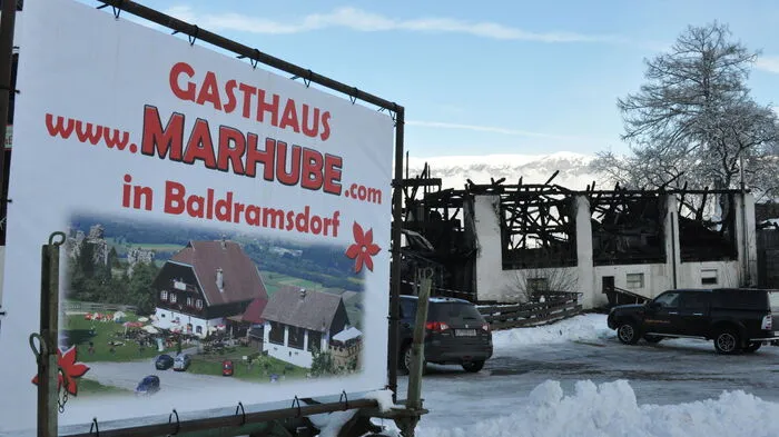 Kontrast: Eine Werbetafel zeigt Marhube samt Stadl, im Hintergrund die Brandruine.