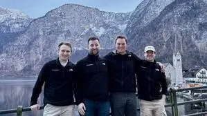 Vier Männer stehen vor einem Zaun, hinter ihnen sind ein See und Berge zu sehen. Sie lachen und haben alle dieselbe, schwarze Jacke an.  | Das Team: Alexander Winter, Ruslan Bakeyev, Daniel Kleinheinz und Maximilian Niedersüß (von links) 