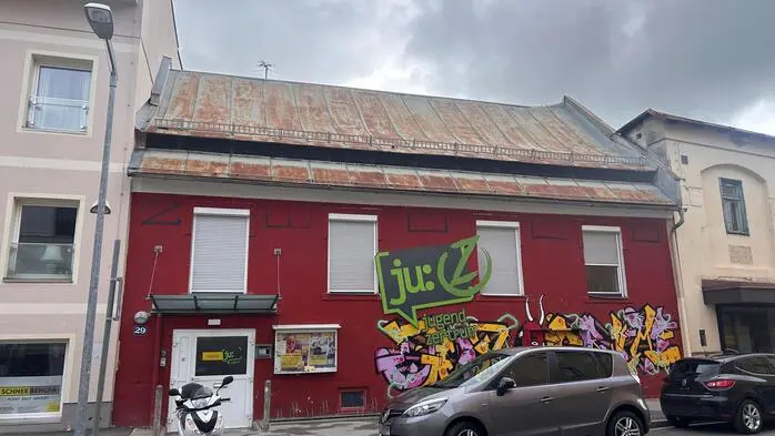 Außenansicht eines roten einstöckigen Gebäudes, davor stehen ein Moped und zwei Autos