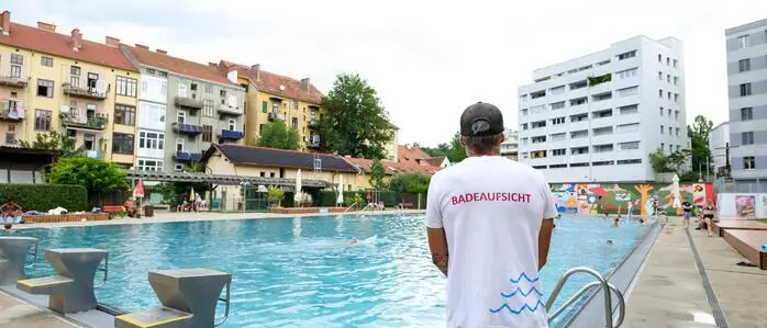 Eine Badeaufsicht steht mit dem Rücken zur Kamera am Rand eines großen Schwimmbeckens im Margaretenbad in Geidorf. Im Hintergrund sieht man die angrenzenden Wohnhäuser.