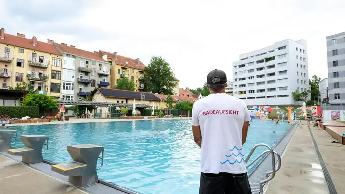Eine Badeaufsicht steht mit dem Rücken zur Kamera am Rand eines großen Schwimmbeckens im Margaretenbad in Geidorf. Im Hintergrund sieht man die angrenzenden Wohnhäuser.