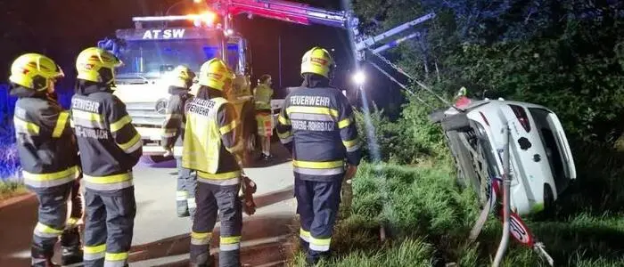 Feuerwehrleute in Uniform im Einsatz, ein Hebekran hebt gerade ein weißes Auto am Straßenrand in die Höhe