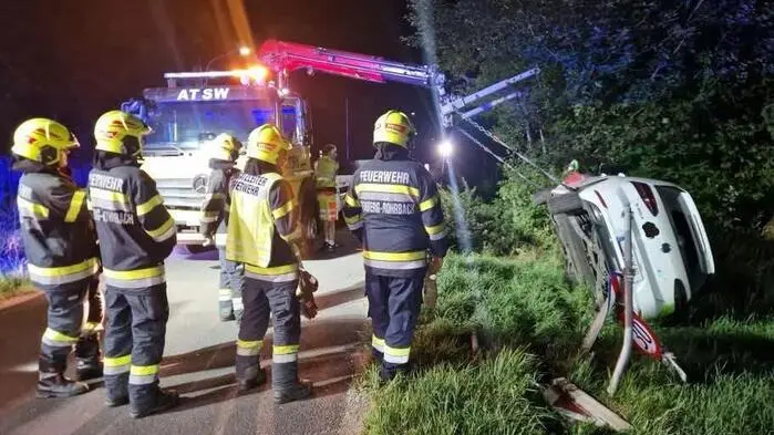 Feuerwehrleute in Uniform im Einsatz, ein Hebekran hebt gerade ein weißes Auto am Straßenrand in die Höhe