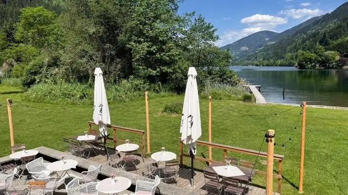 Ein Gastgarten auf einer Wiese, im Hintergrund ein See und Berglandschaft