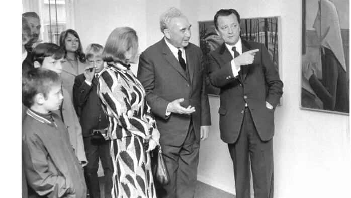 Eine Schwarz-Weiß-Aufnahme von mehreren Menschen, die in einem Museum stehen und ein Bild betrachten. Ein Mann zeigt mit dem Finger auf ein Bild
