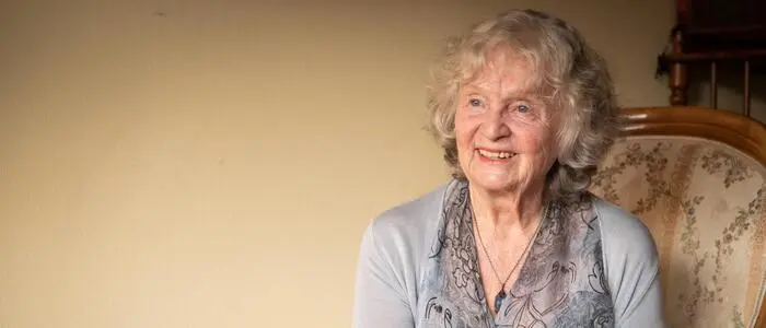 Eine alte Frau mit grauen Locken und einem hellblauen Kleid sitzt auf einem Polstersessel und lächelt.