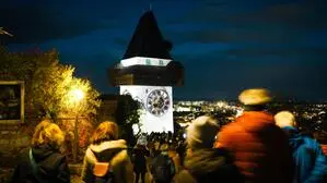 Der Grazer Uhrturm, fotografiert nachts, beleuchtet mit einer Lichtinstallation, rundherum viele Menschen