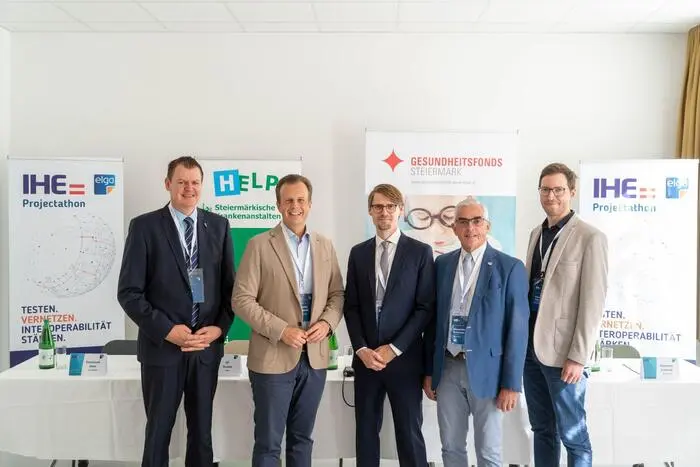 Fünf männliche Personen in Anzügen stehen vor einem Tisch und Plakaten.