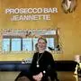 Goldene Wand mit großem Spiegel und der Aufschrift „Prosecco Bar Jeannette“, davor eine blonde Frau mit schwarzem Pullover | Jeannette Staudte eröffnete in Villach ihr erstes Lokal