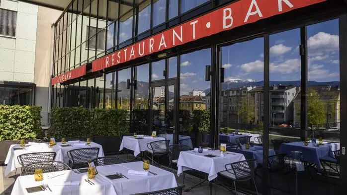 Terrasse eines Restaurants, mehrere Tisch mit weißer Tischdecke, dahinter eine Glasfront mit der Aufschrift „Restuarant, Bar“