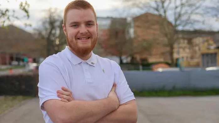 Ein lächelnder Mann mit rotem Bart und weißem Hemd vor einem Wohngebiet