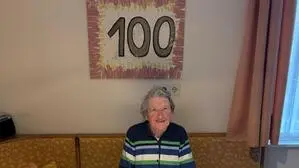 Eine Frau mit grauen Haaren sitzt hinter ihrem Küchentisch, über ihr ist ein Bild auf dem die Zahl 100 steht