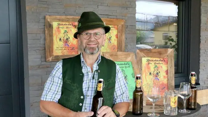 Ein Mann mit Hut hält eine Flasche Bier in den Händen und lächelt in die Kamera.